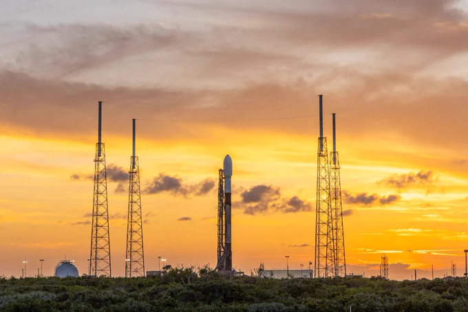 With the sun setting in the background, the SpaceX Falcon 9 rocket sits ready to go surrounded by four towers.