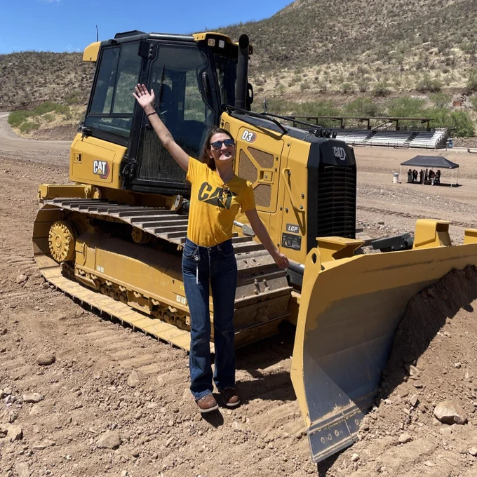 Kasey Spradling holding her her arms out in excitement in front of a CAT construction vehicle. She is also wearing a CAT t-shirt