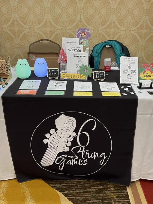 A convention table selling tabletop role playing games with a black banner saying “six string games”