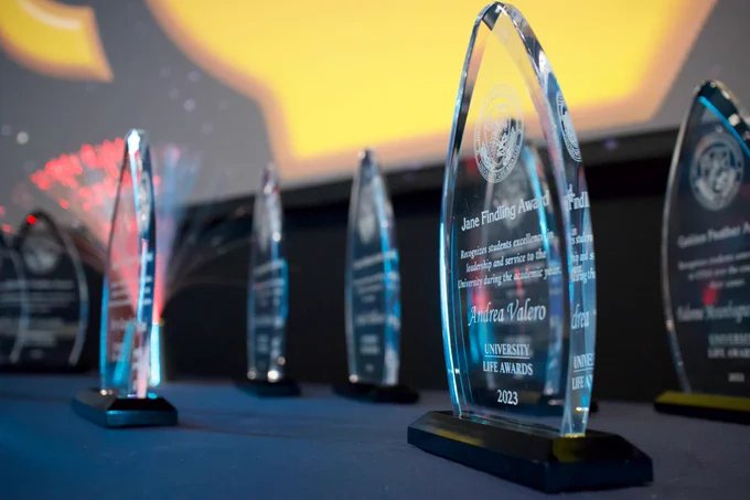 University Life Awards sit on a table with a blue tablecloth on it. The University Life Awards are made out of glass and resemble an oval, with a black stand at the bottom. Awards are etched onto the glass. 