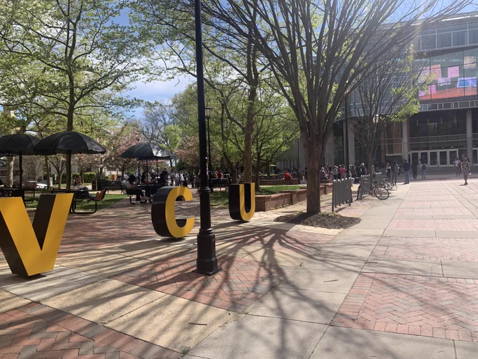 VCU's campus during the spring. Gold VCU sign sits in front of picnic tables. Cabell Library can be seen in the background.