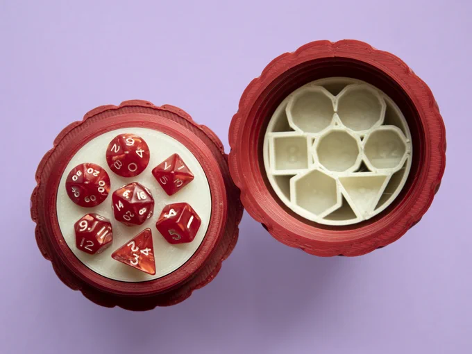 Top view of the two halves of a red egg with a set of 7 mini dice. The top part insert has a set of 7 hollow prisms to hold the dice, and the number of faces of each dice is engraved in the flat base of each prism.