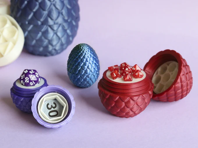Four threaded-inside scaly eggs, or "dragon eggs", either open or closed and in different colors. There's a purple egg open at the front left, with a 30-sided die standing on its bottom half, whilst the top half is facing the viewer, having a hollow prism with a number 30 in it; the prism can hold the die. At the right, there's a similar, bigger, red open egg with a set of 7 mini dice standing on it. Beyond them, there's a closed, smaller blue egg. At the farthest part of the image, left, a much bigger blue egg with half of the insert at its side; both are blurry.