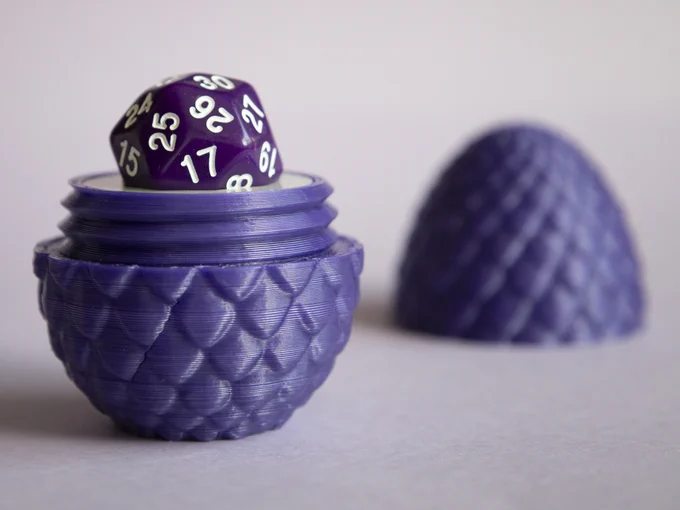 Close view of an open threaded dragon egg with a purple 30-sided die standing on it. Part of the die is below the half egg bottom top line, being held "sinking" in it.