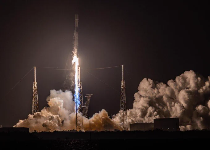 Lift off on the launch pad, surrounded by three towers, at night. Billowing clouds of smoke and bright light.