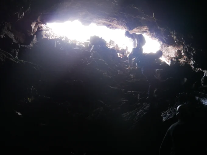 Two silhouettes of scientists are seen as the researchers lower themselves 30 feet through an opening in the ground, which formed where part of a lava tube collapsed. Wearing a headlamp, gloves, and kneed pads, they climb down the tube's rocky walls deep under the ground. Bright sunlight shines at the mouth of the lava tube but only reaches a few feet into the opening, and the scientists lower themselves into the rocky darkness. 