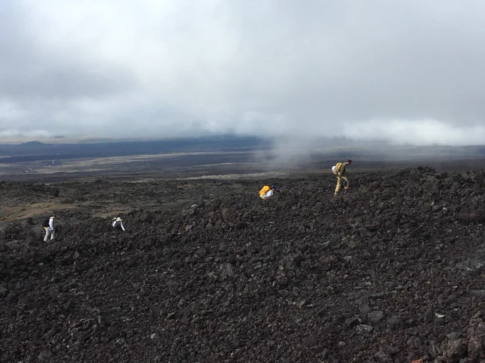 Four scientists hike across the Maua Loa volcano in Hawaii, trekking across dark rubble piles of rocks that form when lava cools quickly. Dense fog almost reaches the ground as they traverse the open landscape. 