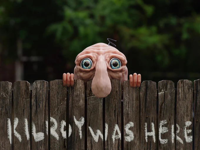 A photo of a lumpy little bald man with a bulbous nose sticking over a wooden fence.  His eyes are big and blue and his tiny fingers are holding him up so just the top half of his head is showing over the fence.  Someone has painted "Kilroy Was Here" underneath him on the fence.
