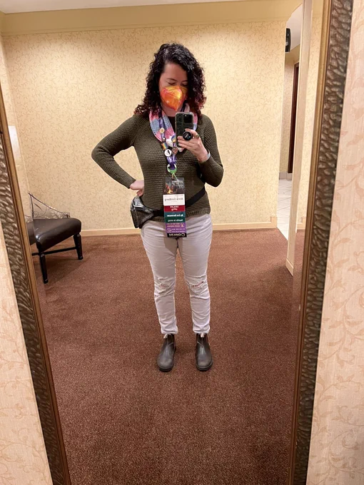Selfie of Anna in a floor length mirror. They’re wearing a trans pride scarf and a convention badge on a lanyard filled with buttons