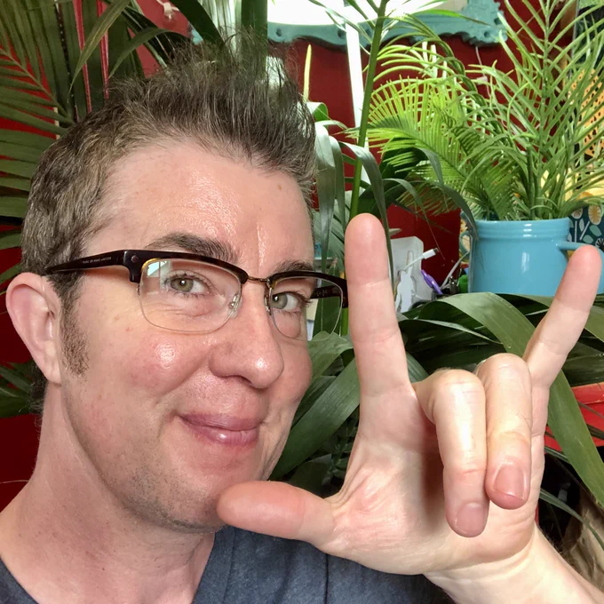 Photo shows Sean signing "I love you," and smiling at the camera. He wears glasses and has short brown hair, wears a blue v-neck tshirt, and the ear hook of his hearing aid can be seen coiling over his right ear.  Behind him are palm trees and other green plants, in front of a dark red wall. 