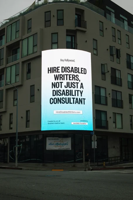 A photograph of a lit up billboard agains the side of a building. It shows bold black text against a blue and white gradient background that reads: Hey Hollywood, Hire Disabled Writers, Not Just A Disability Consultant. 