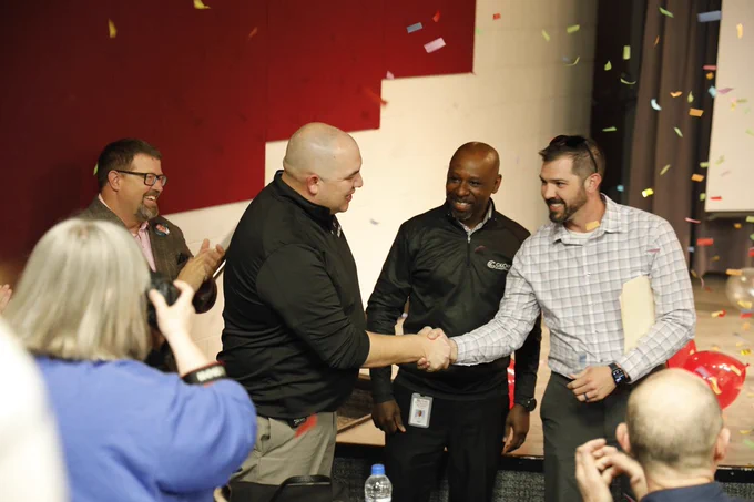 Two men shaking hands while others look on and confetti falls.