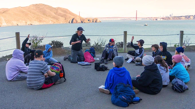 Photo description: A group of young students sits in a circle with their NatureBridge educator on a waterfront bluff. Downtown San Francisco, the Golden Gate Bridge, the Pacific Ocean and a boat are visible in the background.
