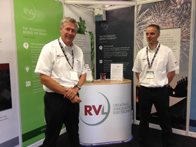 Two electronic engineers in front of a company exhibition stand, with company logo for RVL logo visible, ready to talk to people interested in electronic design or software. The men have friendly approachable expressions. The  background shows exhibition display materials describing the company in more detail, with reference to specific case studies. For example, RVL developed and built the electronics for the London New Year's Eve fireworks display. Another part of the display describes the innovative technology created for hydroponics equipment, so that micro-greens can be grown without human interaction, within a sterile kitchen environment.
