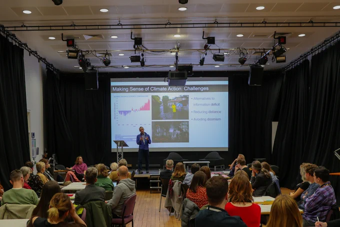 Simon standing at the front of a packed room, with slides behind him saying Making Sense of Climate Action Challenges. 