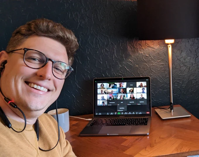 Man with glasses and yellow shirt takes a selfie with a zoom call on a wooden table in the background 