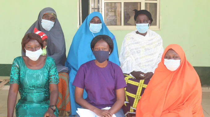 Six women, in two rows of three, are sitting outside. All are wearing face masks. 