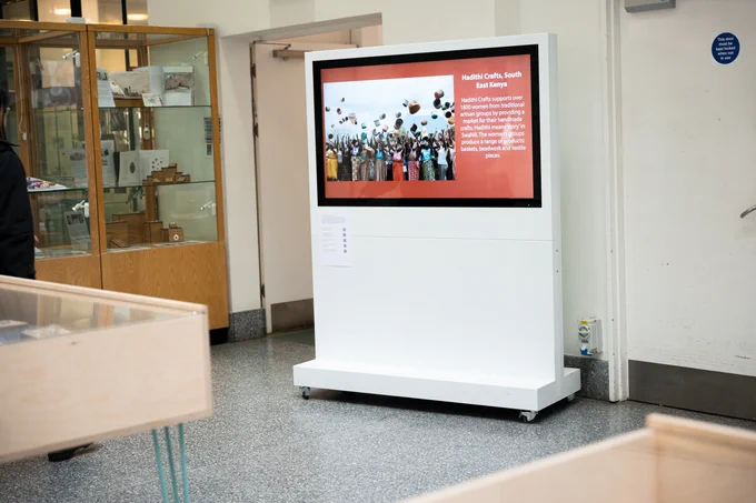 art gallery interior space. a white display with a large screen shows an image of a large group of people throwing baskets into the air. Other wooden display cabinets are visible in the foreground and background. 