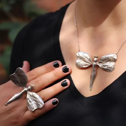 close up image of a silver necklace in the shape of a butterfly worn above a black v neck top. A hand wearing a large silver butterfly ring rests on the chest near the necklace.