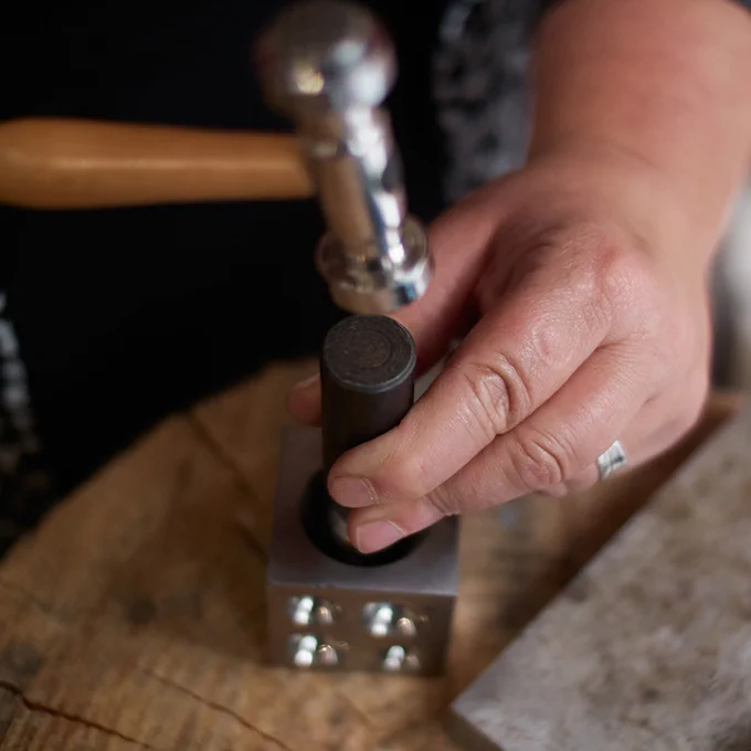 A hammer is visible above a hand holding a component into a metal cube with circular forms on its sides.