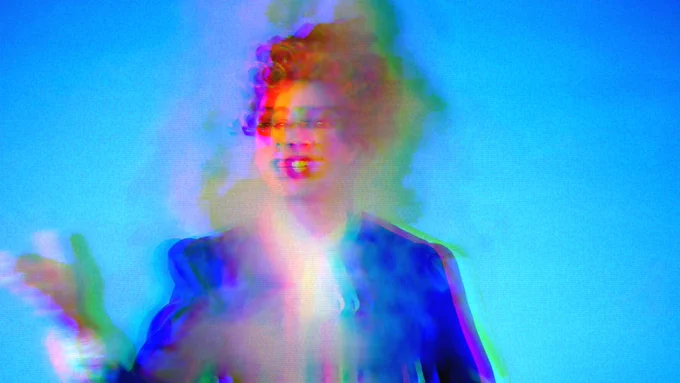 Image is a still from the short film Section 28 and Me by Tom Marshman. Image shows Tom dressed in a Thatcher wig wearing a blue blazer and white blouse with a bow at the neck. They are in the middle of speaking and gesturing with their hand. The image of Tom is blurred by smoke which looks green and white in the light.