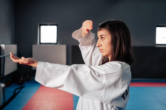 Image is of a woman in a dojo wearing a karate Gi with her arms posed in a defense move.
