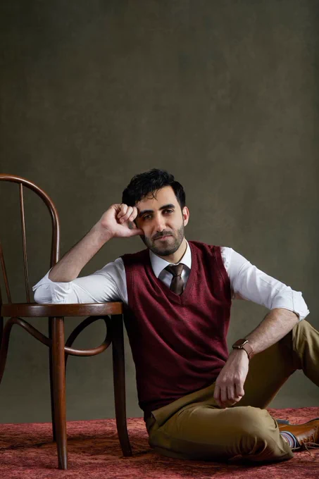 Faran Moradi leans on a wooden chair, in a stylist outfit, against a dark plain backdrop. 