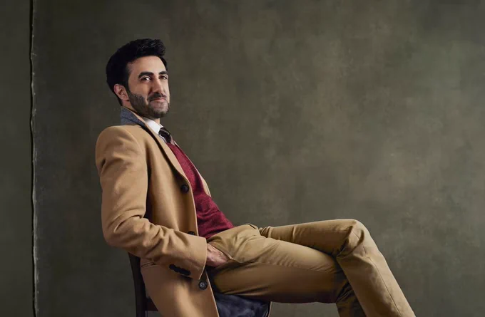 Faran Moradi sits back in a chair, in a tailored tan suit against a dark plain backdrop. 