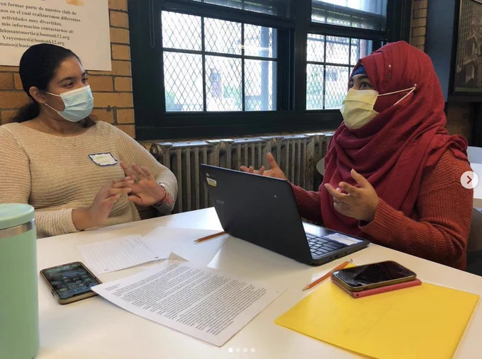 A tutor on the left talks to a student on the right. They are sitting at a table next to a laptop and papers.