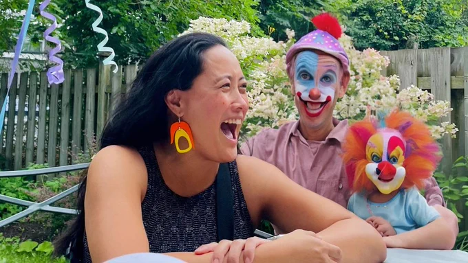 A woman, mid-laugh, looks off to the side. She's sitting at a table in a backyard. Beside her are two bizzare-looking AI-generated clowns. One looks like a daddy clown with a wide grin and a shoddy face-paint job. His hat looks like a rainbow, polka-dotted paperboy cap with a pom pom. He wears a collared shirt that also looks like a golf shirt. He's holding a little clown child with warped facial features and wide eyes. Her hair looks like it's made out of tutu skirts. 