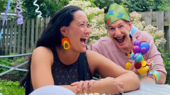 Woman laughing with an older person with an unnaturally large and angular mouth and slouching neck that seems to melt into their backwards-looking shirt. She’s looking off to the side but he is starting straight at her. Random small balloons and a green hat with stars make this one a clown? He wears no clown makeup
