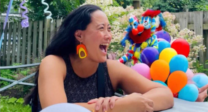 Woman laughing next to garbled clown with two noses and no discernible head, behind a bunch of balloons