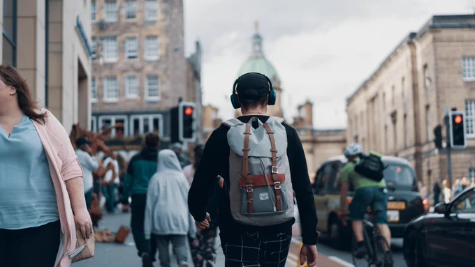 A man wearing dark clothing, a black cap, grey backpack and headphones walks along a busy city street with people walking on his left, cars waiting at a red light and a cyclist passing on his right.