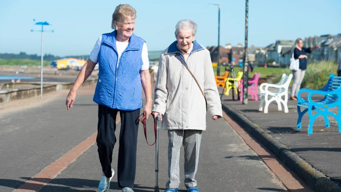 A lady with a blue embroidered gilet and dark trousers smiles as she walks alongside another lady in light grey trousers, jacket and walking stick as they move along a promenade. 