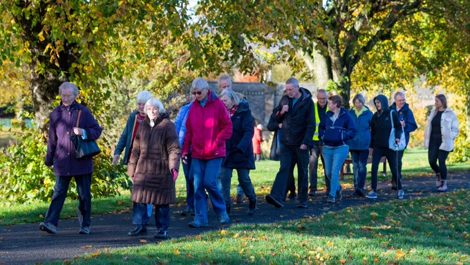 A large group of people in outdoor clothing and sunglasses enjoy a walk along a path through an urban park, with trees illuminated by the sun behind them.