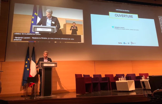 Discours au pupitre de Benjamin Laurent a l’ouverture de la soirée. Derrière lui les drapeaux français et européen. Au dessus un écran avec la velotypie.