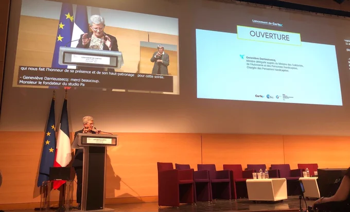 Discours au pupitre de la@ministre a l’ouverture de la soirée. Derrière elle les drapeaux français et européen. Au dessus un écran avec la velotypie.