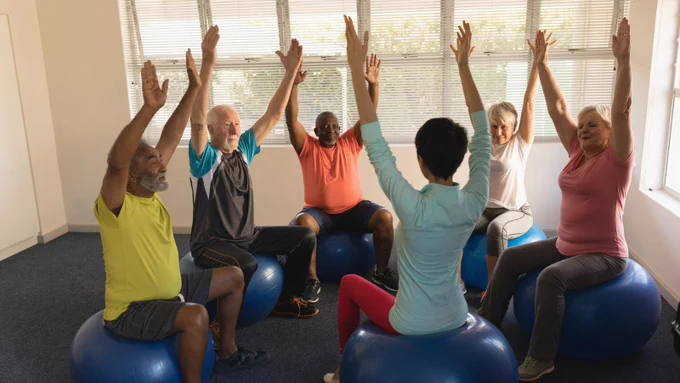 Photo of 6 adults sitting on blue exercise balls with their hands in the air. 5 are sat in an arc facing the instructor