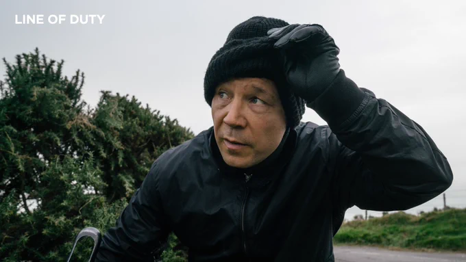 As John Corbett in Line of Duty. He lifts a black balaclava over his face while looking off camera to his right.