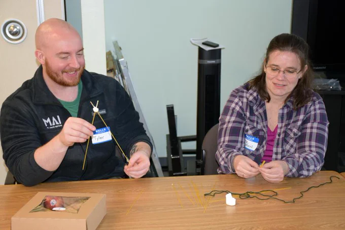 Two people sit at a table trying to figure out how to create a tall structure using only raw spaghetti noodles, string, tape, and a marshmallow.