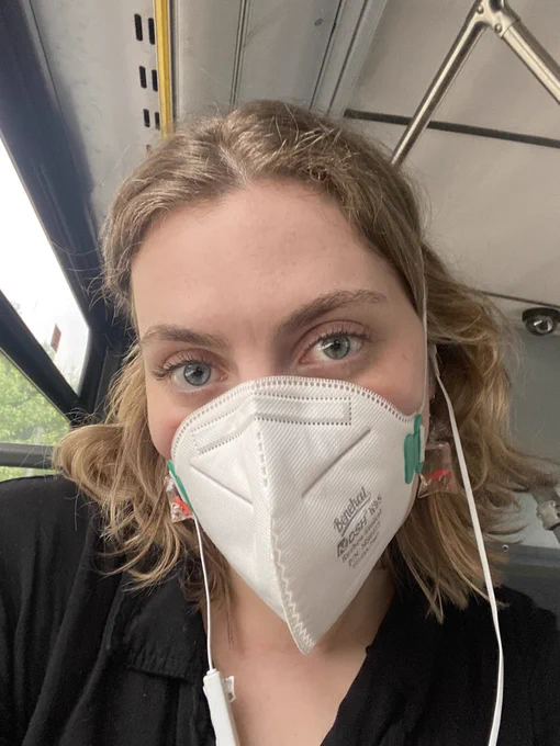 Selfie of a young white woman with shoulder length blonde wavy hair and blue eyes. She’s wearing a white N95 and corded white earbuds.