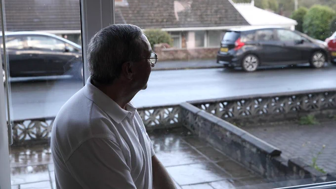 Man in a white shirt looking out from his living room window towards the street ahead, where there are parked cars. 