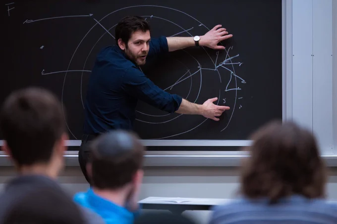 Man in blue shirt points to drawing on chalkboard as students look on