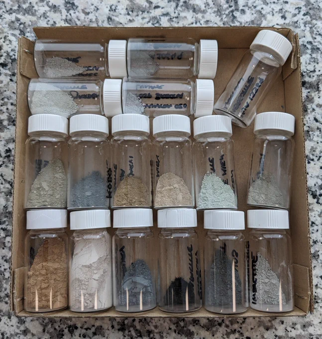 A brown cardboard tray with 17 glass sample vials that have white lids. Each vial has a powdered sample in it, of varying colours. Some are grey, white, orange, brown, black... Some are almost full and some are almost empty.