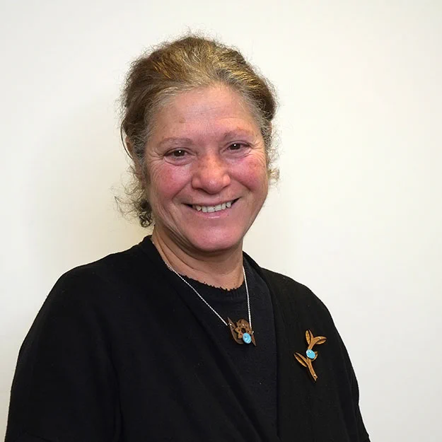 a woman in a black top and cardigan wears a wood and enamel brooch resembling an olive branch pinned to their chest and a wood and enamel pendant around their neck resembling a posy of flowers.
