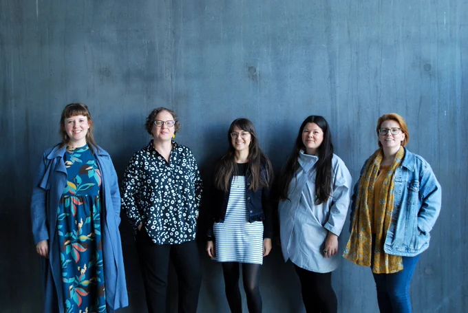 Photograph of five women standing against a blue-grey concrete wall smiling at the camera. They pictured three quarter length, and are wearing clothes in blue, black and white tones.