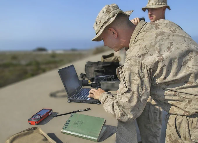 A Marine in camouflage types on a laptop on a concrete wall, while another Marine with binoculars looks in the distance. There is a GPS device, a notebook, and a satchel in the foreground. (Photo Credit: Cpl. Miguel Rosales, U.S. Marines Corps; modified by the Army Geospatial Center)
