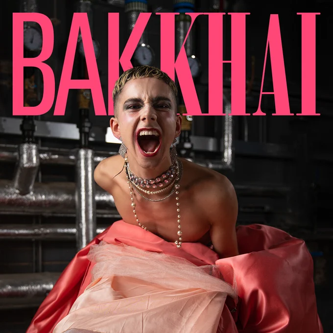 BAKKHAI in classical pink text behind a photo of a performer wearing a pink skirt and lots of necklaces screaming at the camera  