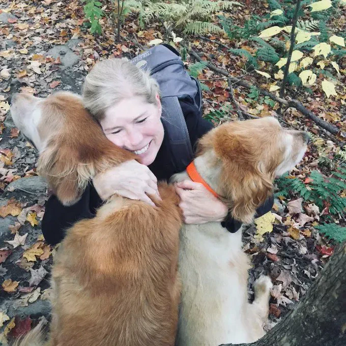 Two golden retrievers (Baylor and Sampson) being hugged by Aynsley Floyd, a woman with blond hair. Aynsley is kneeling in front of them, with her arms around them, smiling with her eyes closed. She's wearing a backpack and they appear to be on a hiking trail in the woods.