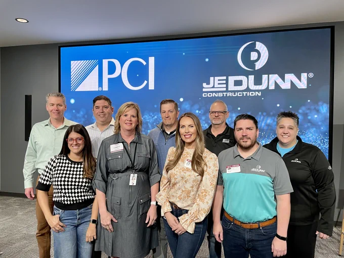 A group of people pose for the camera in front of a screen with PCI and JE Dunn's logos on them. 